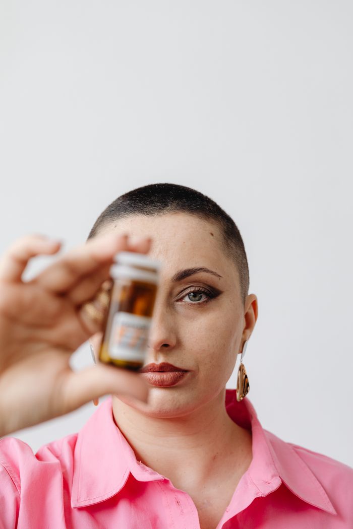 A bald woman in a pink top holds a medicine bottle, focused in a studio shot.
